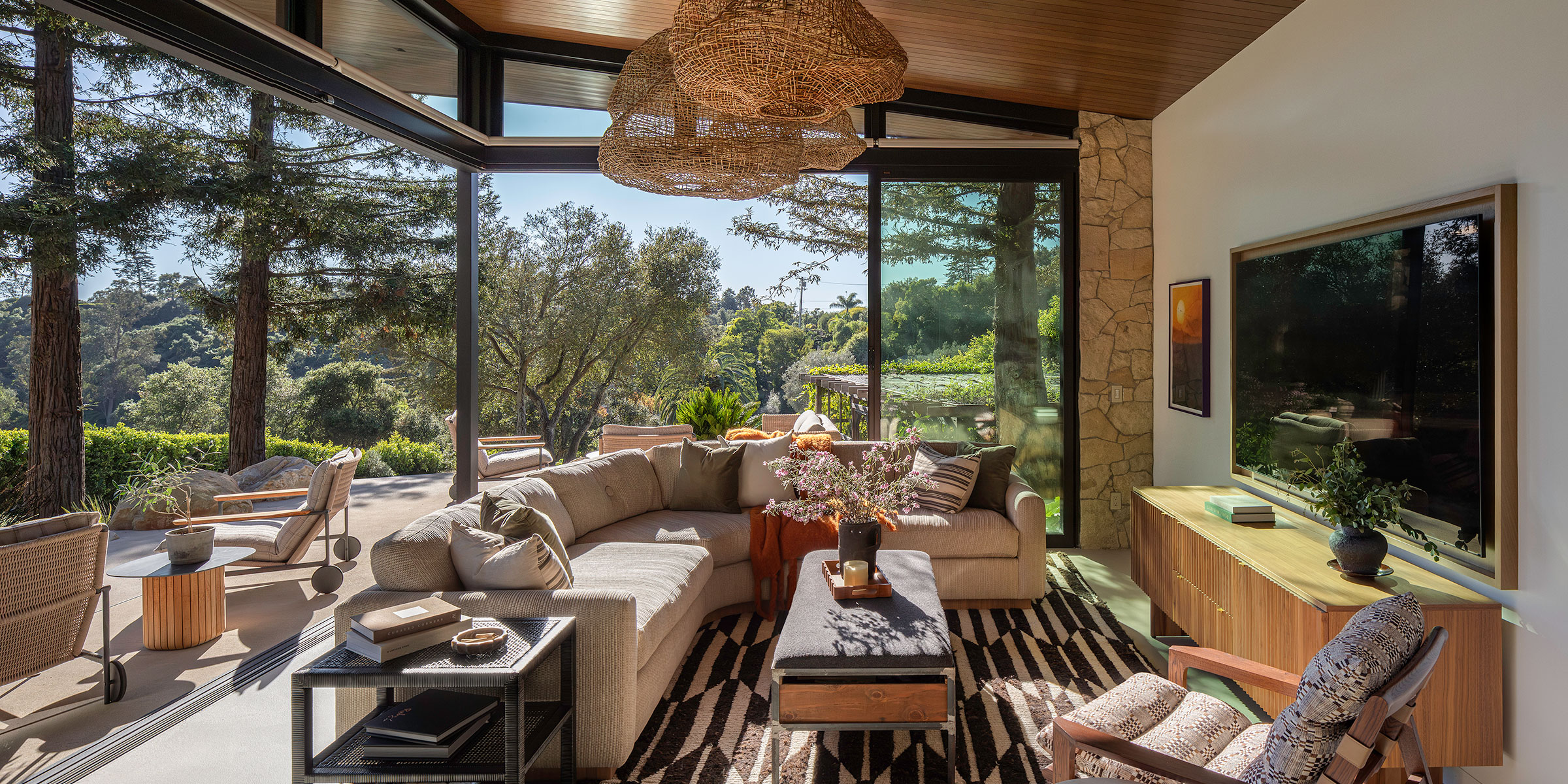 A modern living room with large glass walls showcases a lush, green outdoor view. With natural wood furniture and woven light fixtures, it beautifully blends indoor and outdoor vibes—ideal for interior design services in Montecito and the Central Coast.