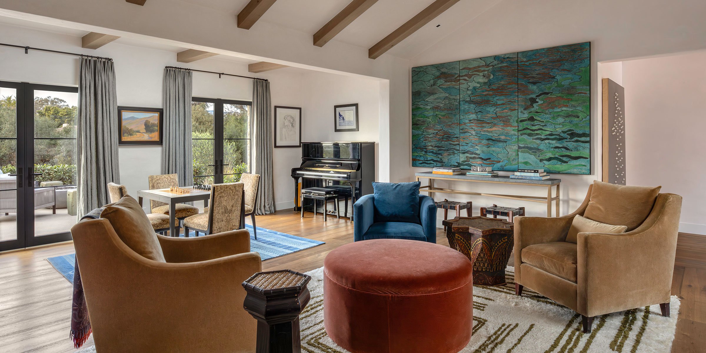 A stylish living room, designed with Santa Barbara interior design services, features tan and blue armchairs, a round red ottoman, piano, dining table, abstract artwork, and French doors welcoming natural light.