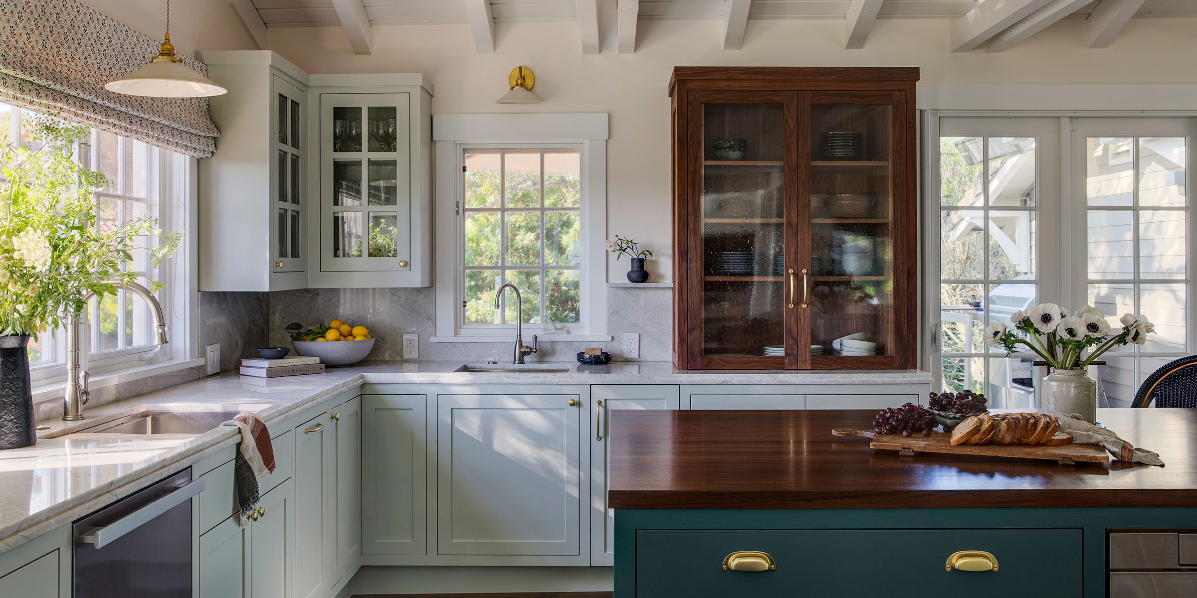 Bright kitchen with white cabinets, a wooden island countertop, glass-front cupboards, and fresh flowers. Sunlight streams in as fruit and bread are arranged on the counters—perfect inspiration for Interior Design services for Santa Barbara and Montecito.
