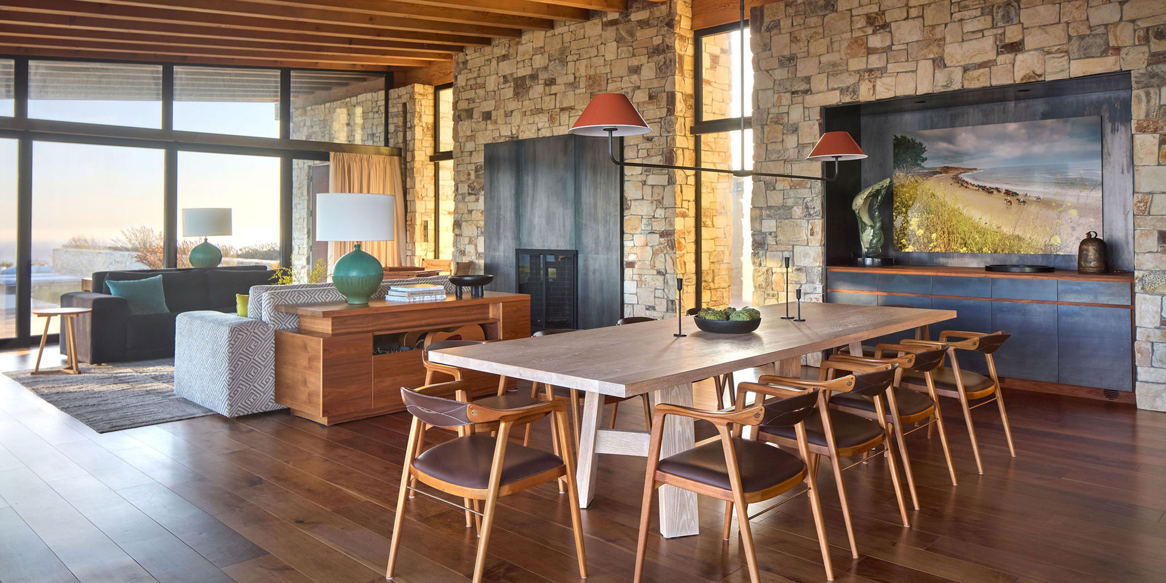 Modern dining and living area with stone walls, large windows, wooden furniture, and a scenic landscape artwork. Natural light fills the room—perfect inspiration for Interior Design services in Santa Barbara, Montecito, and the Central Coast.