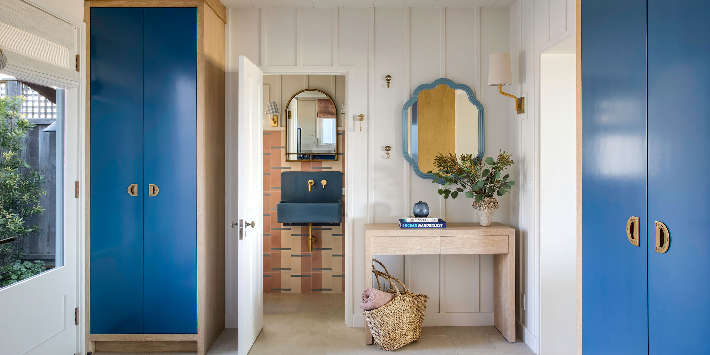 A bright entryway with blue cabinets, a wooden console table with a plant and books, a wicker basket, and a mirror; an open door reveals a bathroom with a blue vanity—showcasing stylish interior design services for Santa Barbara and Montecito.