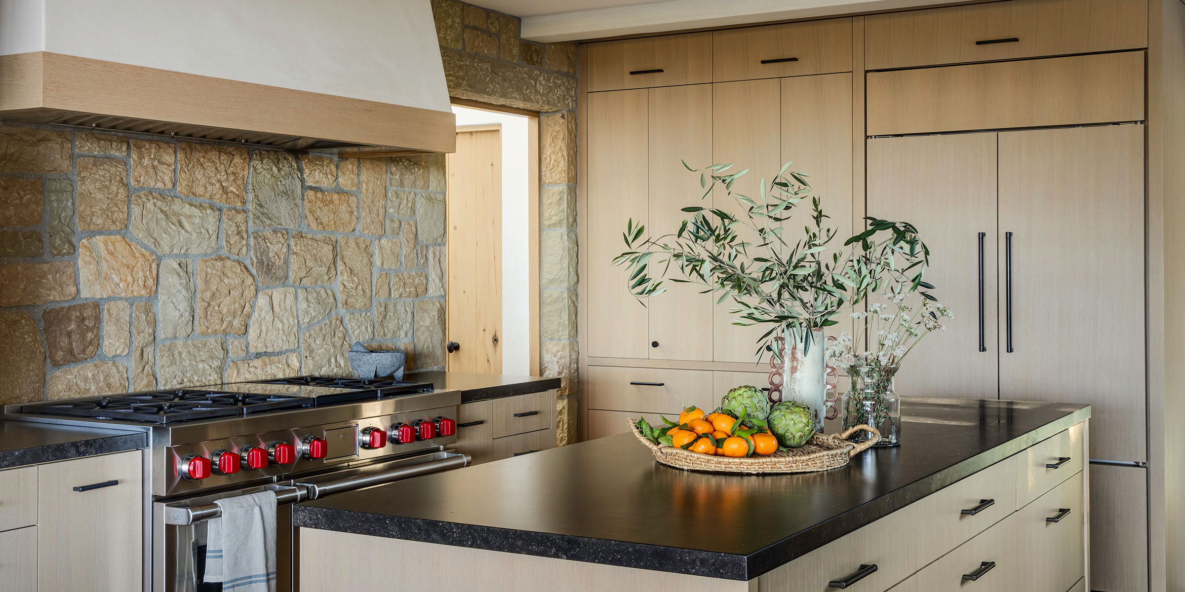 Modern kitchen with light wood cabinets, stone backsplash, a stainless steel stove with red knobs, and a black countertop island decorated with a glass vase of greenery—ideal for those seeking interior design services in Santa Barbara and Montecito.
