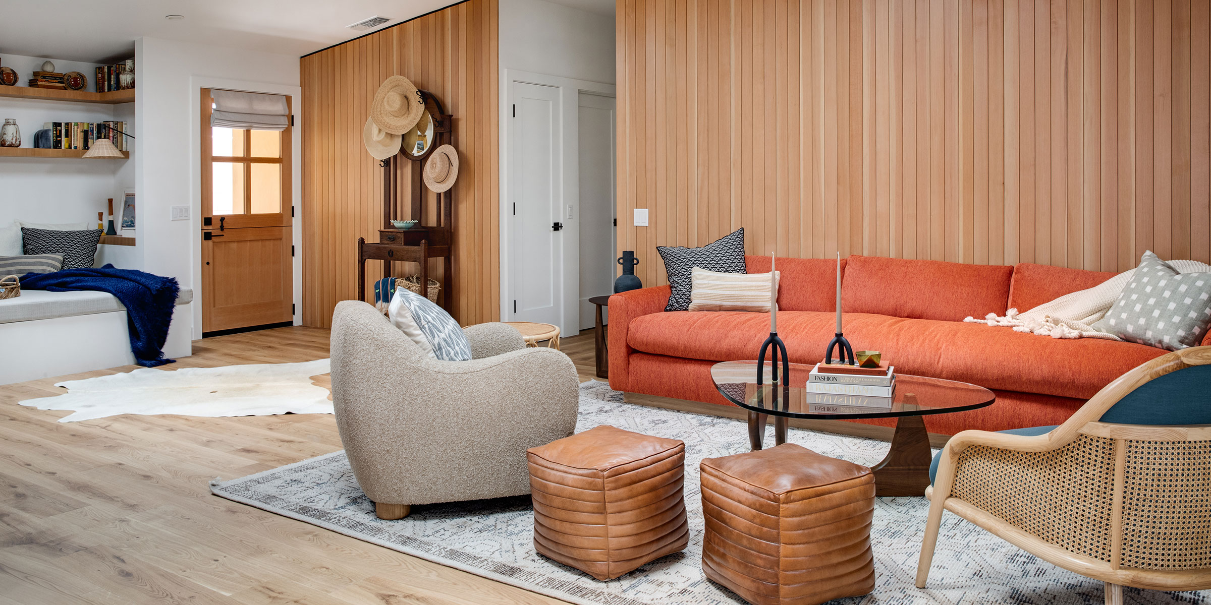 A cozy living room in Santa Barbara style, featuring a red sofa, beige armchairs, leather poufs, a glass coffee table, wood-paneled walls, and hats near the door—perfect inspiration for Interior Design services for Montecito and the Central Coast.