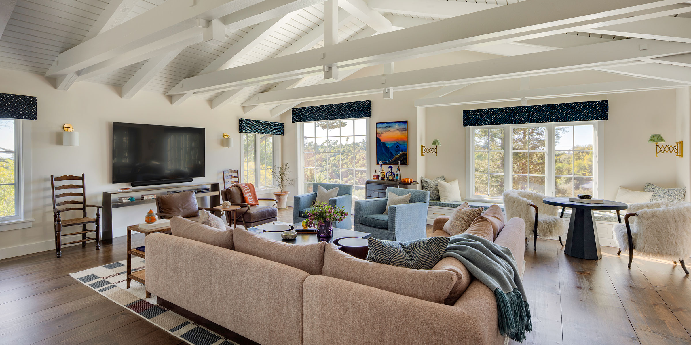 Spacious, bright living room with exposed white beams, large beige sectional sofa, armchairs, flat-screen TV, wood floors, large windows, and modern decor with colorful accents.