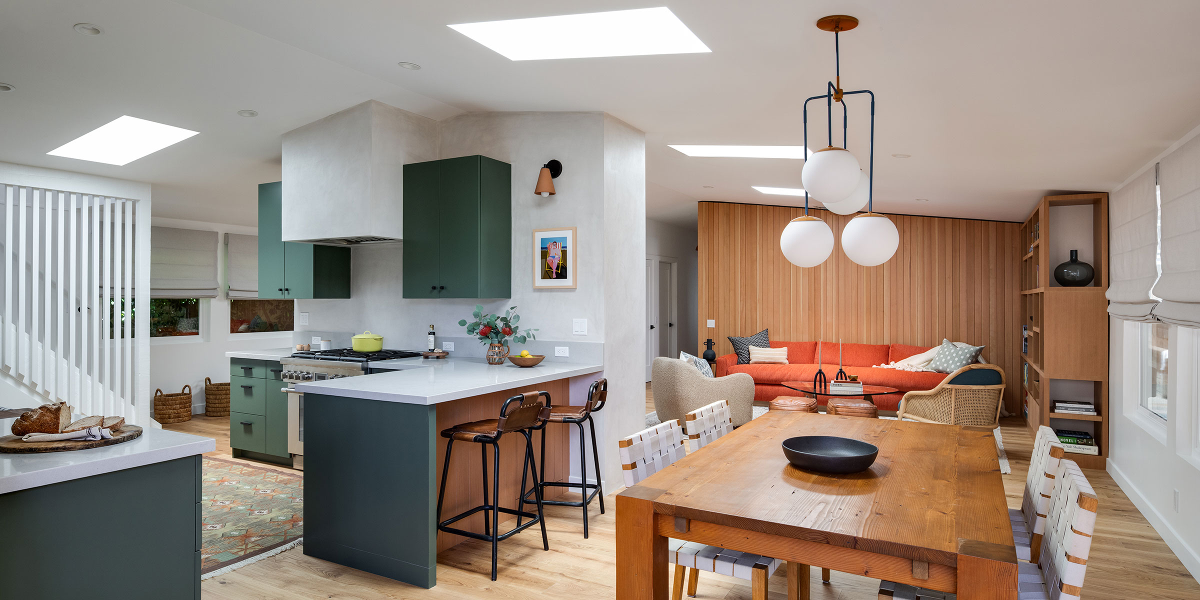 Open-concept kitchen and living area with green cabinets, wood accents, a wooden dining table, modern pendant lights, and an orange sofa against a wood-paneled wall. Natural light streams through skylights—Interior Design services for Santa Barbara.