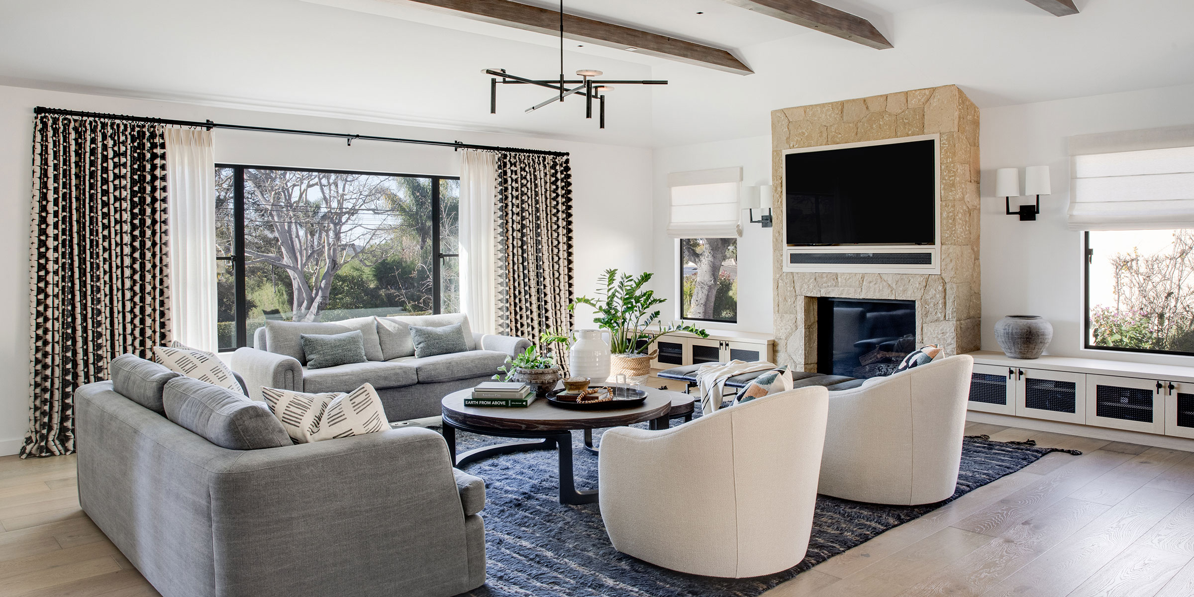 Bright living room with large windows, gray sofas, two white armchairs, a stone fireplace with a mounted TV, patterned curtains, and modern accents—expertly styled by Interior Design services for Santa Barbara and Montecito.