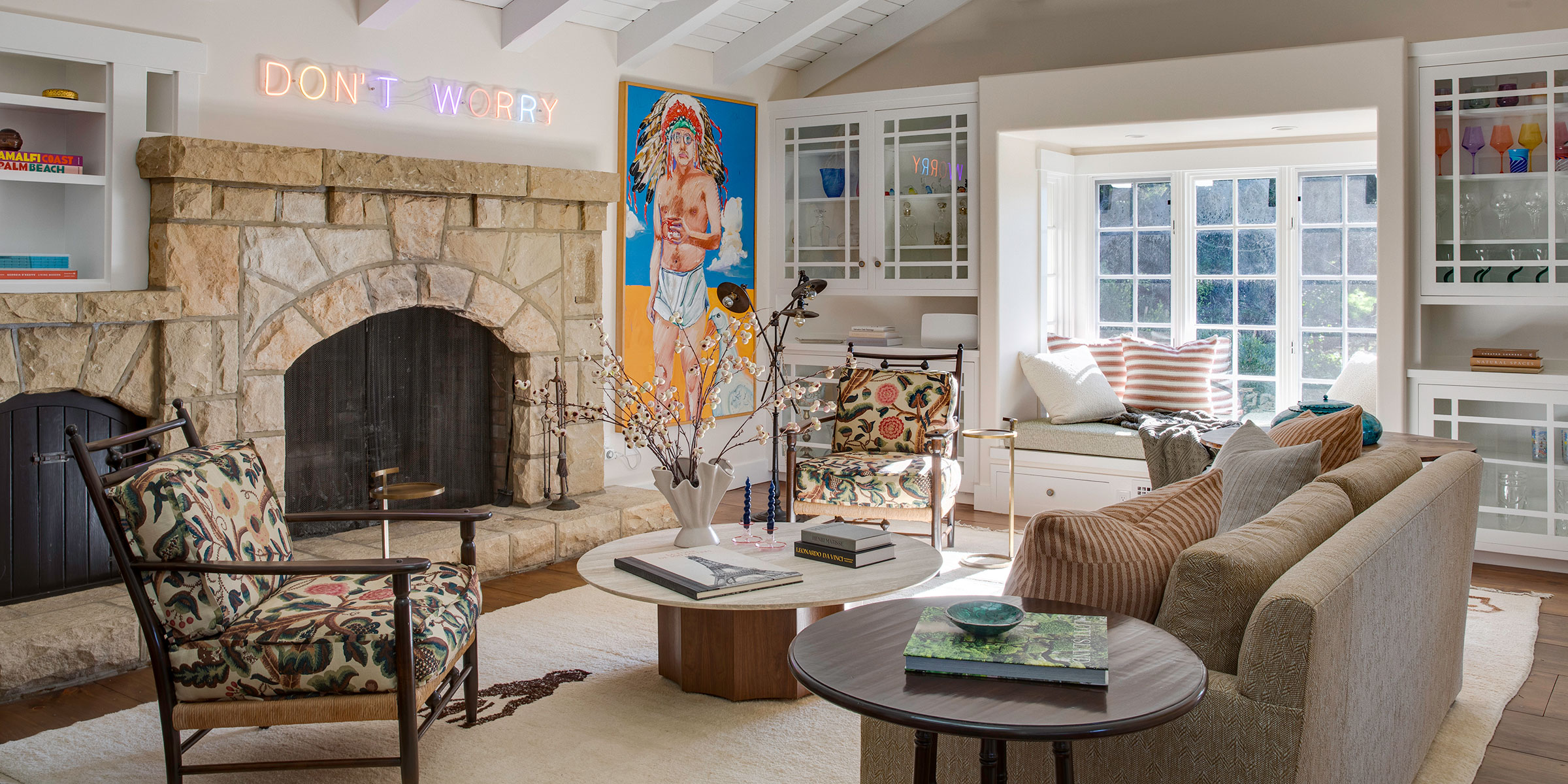 A cozy, sunlit living room with a stone fireplace, eclectic furniture, vibrant painting, bookshelves, and a “DON’T WORRY” neon sign—perfect inspiration for interior design services in Montecito and the Central Coast.