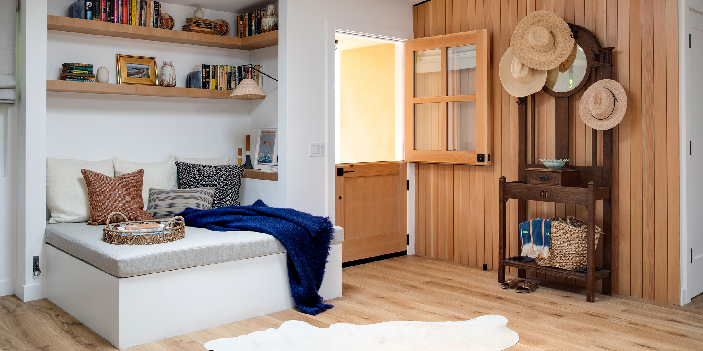 A cozy, modern entryway features light wood floors, a reading nook with pillows and a navy throw, open shelves with books and decor, and a vintage hall tree. Interior Design services for Santa Barbara Montecito & beyond can bring this vision to life.