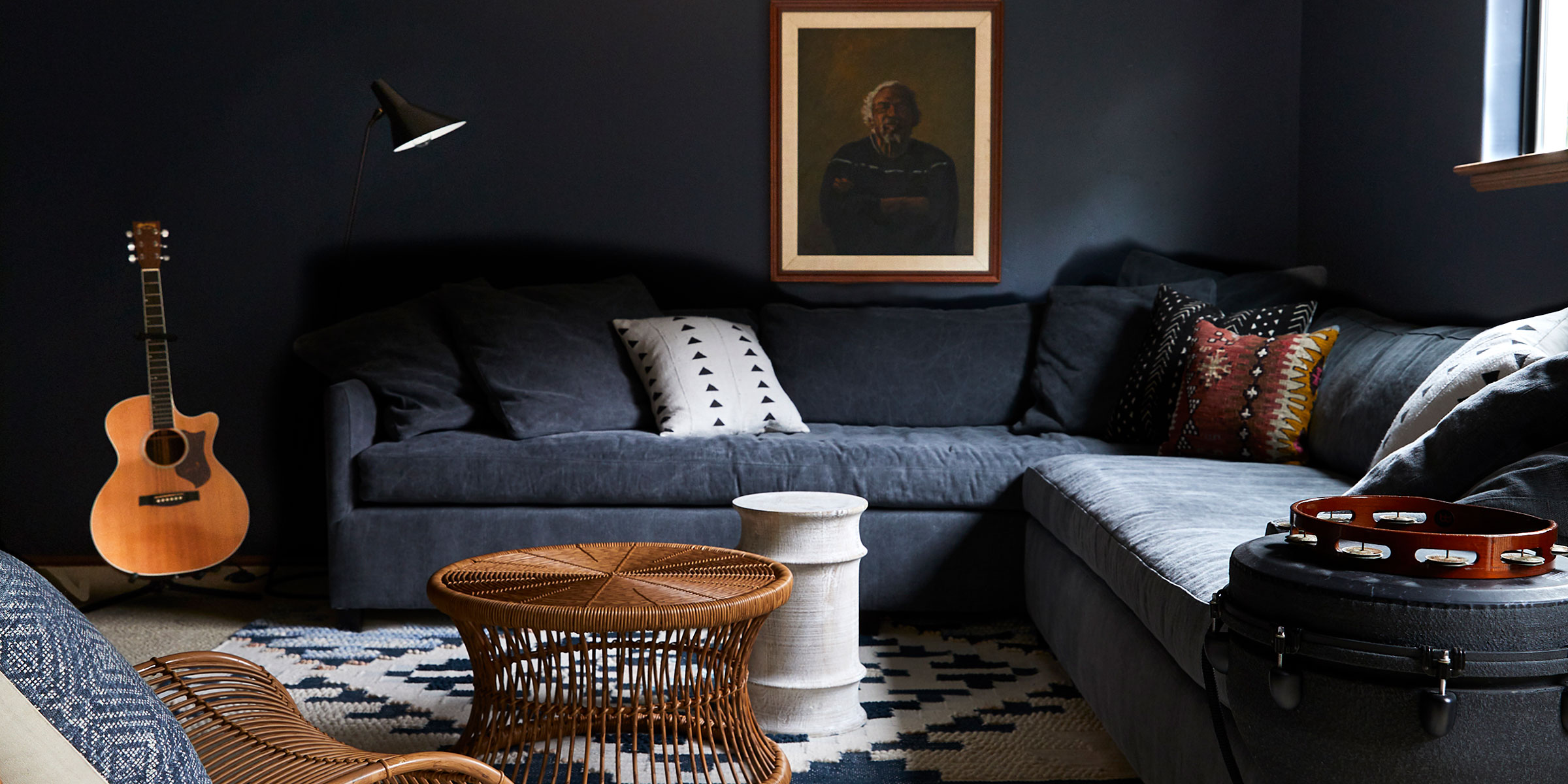 A cozy living room in Montecito features a dark sectional sofa, patterned pillows, a wicker coffee table, and musical touches. Natural light pours in—perfect inspiration for Interior Design services across Santa Barbara and the Central Coast.