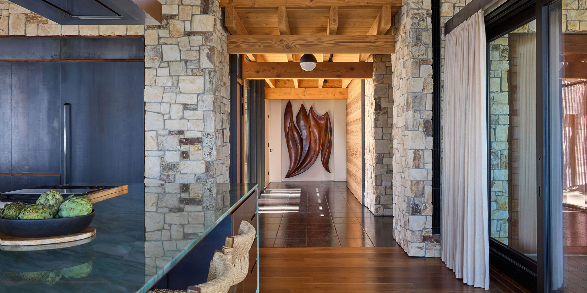 Modern interior with stone walls, wood beams, and a glass countertop. A hallway with dark flooring leads to a wall-mounted abstract wooden sculpture at the far end, and light curtains hang to the right.