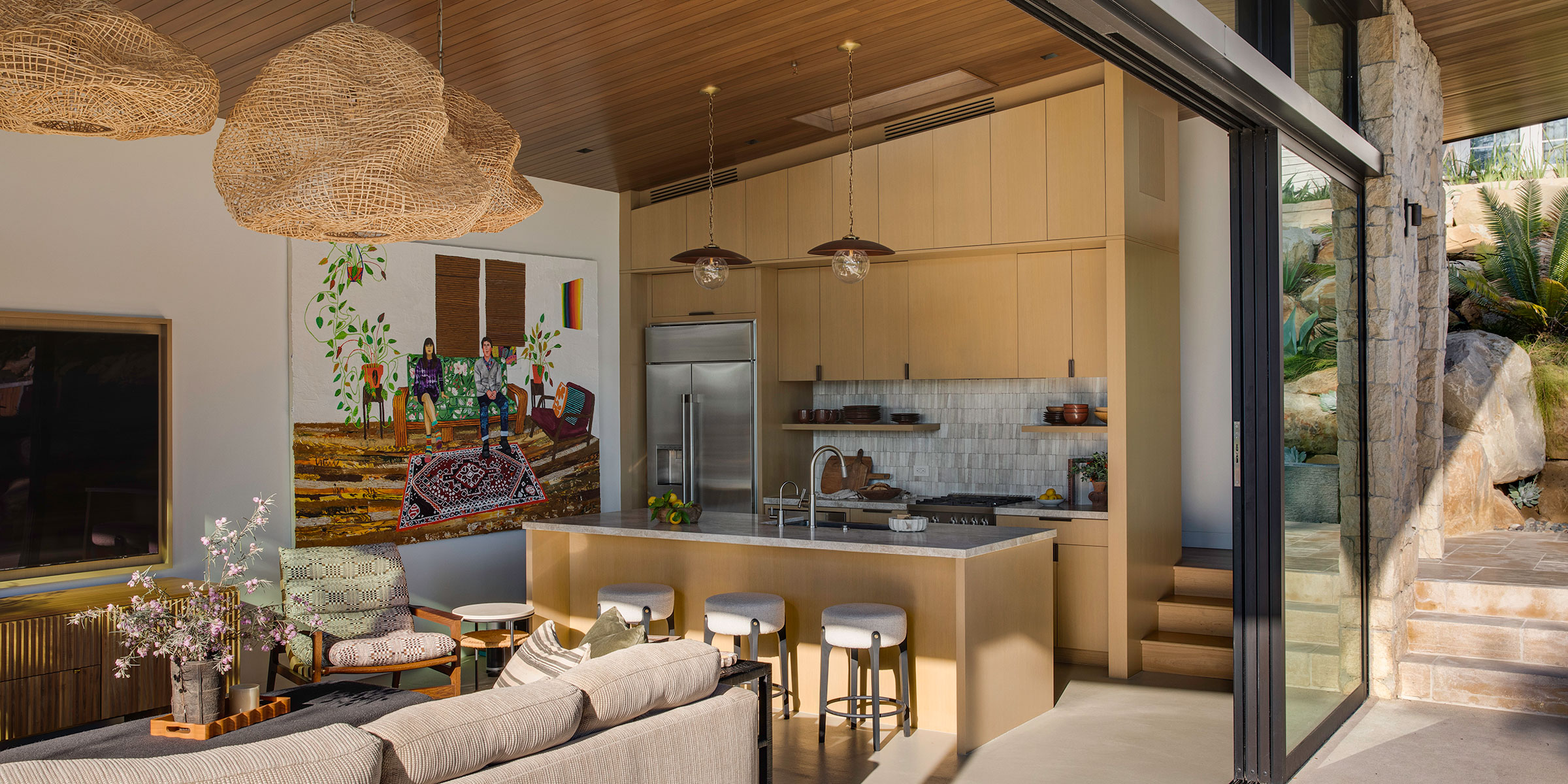 Open-concept kitchen and living area with light wood cabinets, a center island, modern appliances, woven pendant lights, wall art, and large glass doors to a stone staircase—reflecting refined Interior Design services for Montecito and the Central Coast.