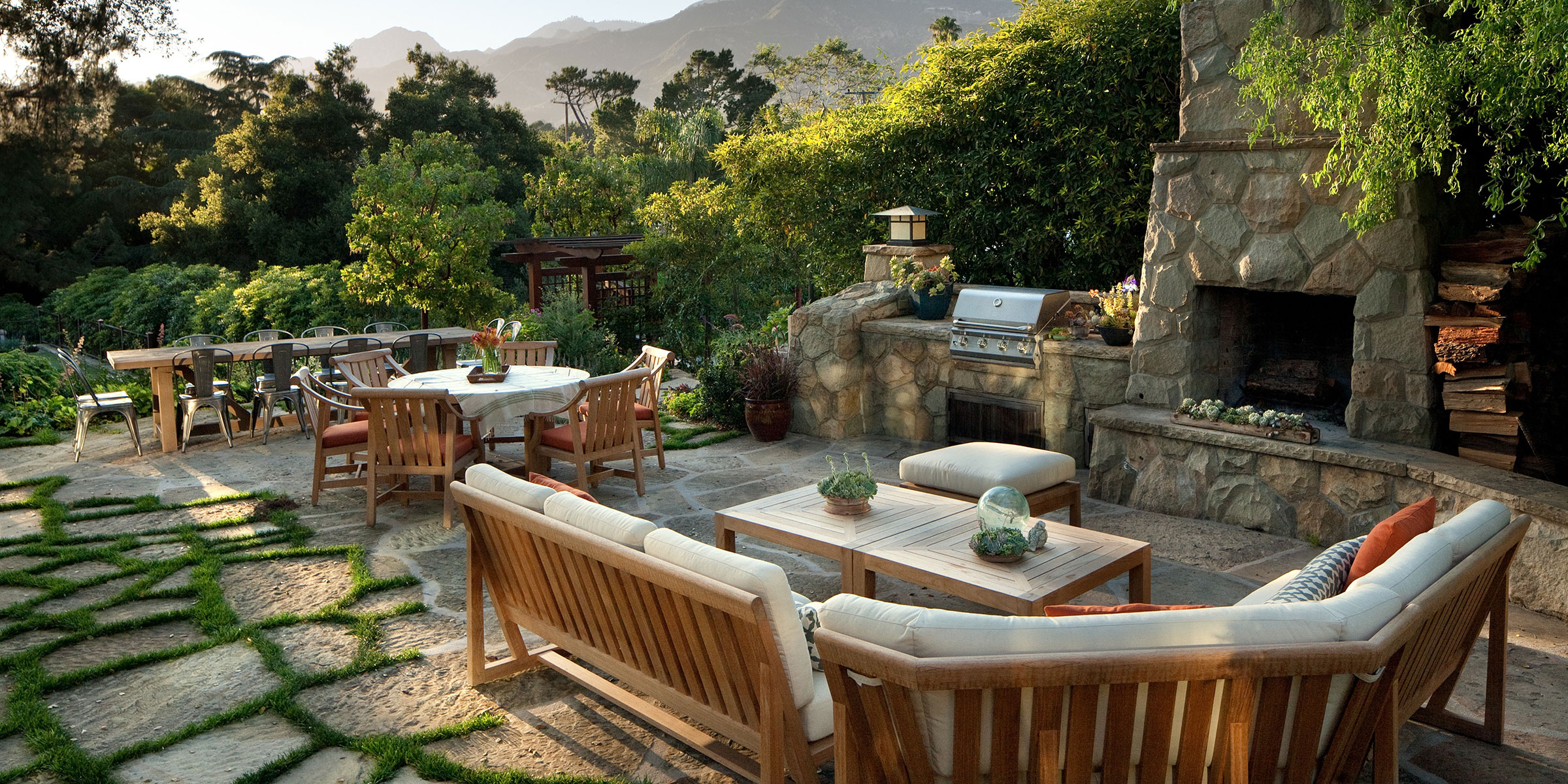 A spacious outdoor patio with wooden furniture, a dining table, a grill, and a stone fireplace overlooks lush greenery and mountains—perfect inspiration for interior design services in Santa Barbara, Montecito, and the Central Coast.