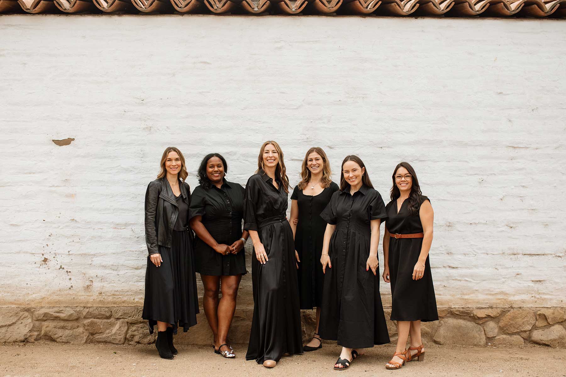 JRSID-team-photo-2026 JRSID team photo. Six women stand side by side in front of a white brick wall with a tile roof, all wearing black dresses and smiling at the camera.